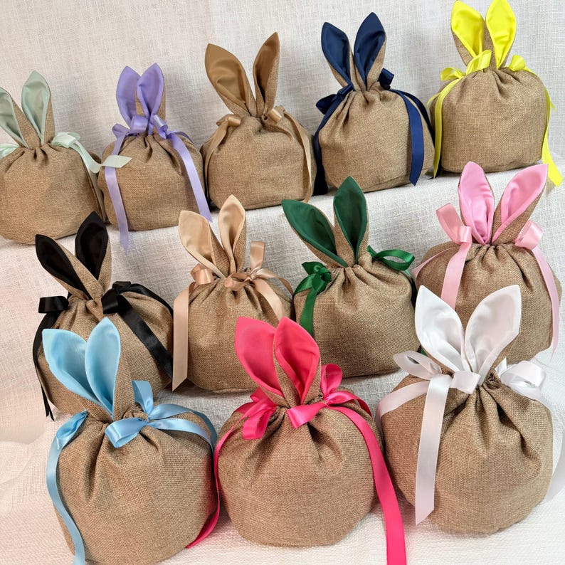 May include: A collection of burlap gift bags, each adorned with bunny ears in various colors, including blue, pink, green, and yellow. The bags are tied with matching satin ribbons, creating a festive and charming presentation.
