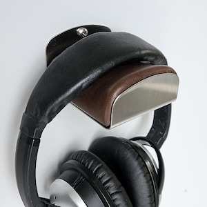 May include: A wall-mounted headphone stand featuring a brushed metal bracket with a brown leather-like padded rest. A pair of black and silver over-ear headphones with black ear cushions is hanging securely from the stand against a white wall.