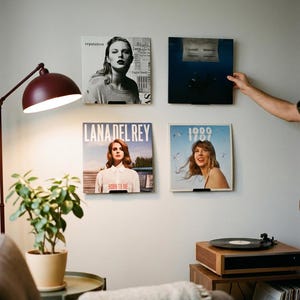 May include: Four square vinyl record covers displayed on a wall, including Taylor Swift's Reputation and 1989, and Lana Del Rey's Born to Die. The covers are mounted on small black wall brackets, arranged in a grid pattern above a wooden record player.