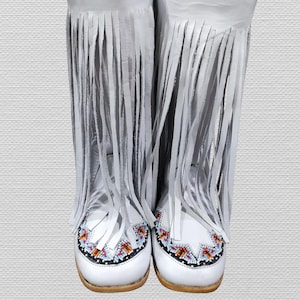 Könnte beinhalten: Ein Paar weiße Lederstiefel mit langen Fransen. Die Stiefel haben ein Perlendesign in Rot-, Orange-, Blau- und Schwarztönen im Zehenbereich. Die Stiefel haben eine hellfarbige Sohle.