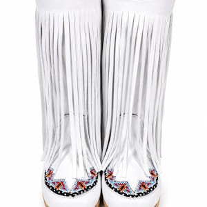 Könnte beinhalten: Ein Paar wadenhohe weiße Stiefel mit langen, vertikalen Lederfransen am Schaft. Der Zehenbereich ist mit einem geometrischen Muster aus kleinen, mehrfarbigen Perlen in den Farben Blau, Orange, Rot und Schwarz verziert.