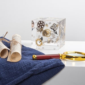 Puede incluir: Un cubo transparente lleno de varios engranajes y piezas de relojería dorados y plateados. El cubo está sobre una superficie de tela azul junto a una lupa con marco dorado.