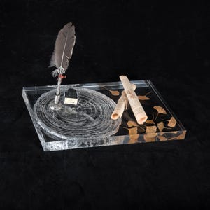 May include: A clear acrylic desk tray with a decorative wood slice base. A silver quill pen with a grey feather, ink bottle, and rolled paper scrolls are on the tray. The tray's right side has gold leaf accents.