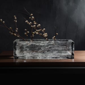 May include: A clear glass vase with a textured surface, filled with dried branches with small white flowers. The vase is sitting on a wooden surface.