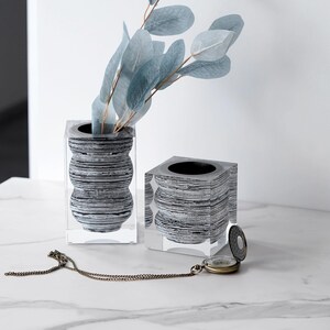 May include: Two decorative vases with a clear, cube-shaped exterior and a gray and black striped interior. One vase holds blue-green leafy branches. A vintage pocket watch with a chain rests nearby. The vases are on a white marble surface.