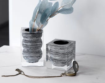 Puede incluir: Dos jarrones decorativos con un exterior transparente en forma de cubo y un interior a rayas grises y negras. Un jarrón contiene ramas de hojas azul verdoso. Un reloj de bolsillo vintage con cadena descansa cerca.