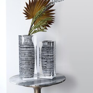 Puede incluir: Dos jarrones decorativos con un patrón de rayas blancas y negras, colocados sobre una mesa con tapa de mármol. Los jarrones son rectangulares con lados de acrílico transparente. Un jarrón contiene plantas secas con hojas marrones y verdes.