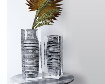 Op de afbeelding: Twee decoratieve vazen met een zwart-wit streepjespatroon, op een tafel met marmeren blad. De vazen zijn rechthoekig met heldere acryl zijkanten. Eén vaas bevat gedroogde planten met bruine en groene bladeren.