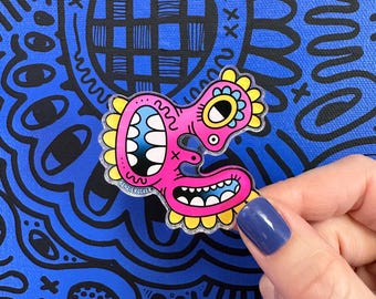May include: A pink, cartoonish enamel pin with a wide open mouth and large teeth. The pin has yellow and blue accents and is held against a blue background with black doodles.