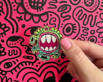 May include: A colorful enamel pin featuring a cartoon monster with a wide open mouth, green body, and pink teeth. The pin is held against a vibrant pink background with black doodle art.