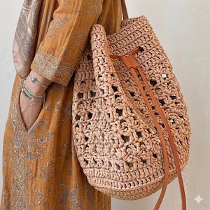 Könnte beinhalten: Beigefarbene Korbtasche mit offenem Webmuster. Sie hat einen Kordelzugverschluss und braune Lederriemen. Die Tasche wird von einer Person getragen, die ein langes, gemustertes Kleid trägt.
