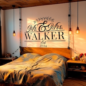 A stylish vinyl decal on a wall above a bed, featuring elegant script that reads 'Mr. & Mrs. WALKER' with 'Est. 2024' below. The design is black against a light wall, complementing the cozy bedroom decor.