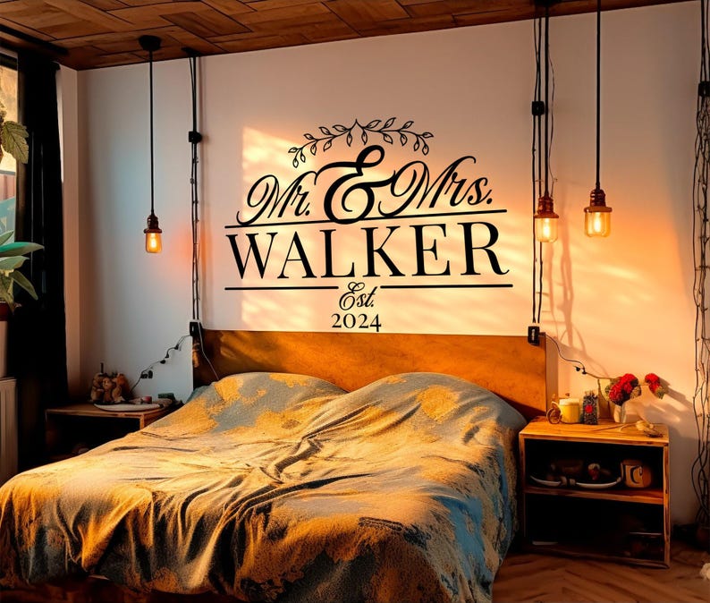 A stylish vinyl decal on a wall above a bed, featuring elegant script that reads 'Mr. & Mrs. WALKER' with 'Est. 2024' below. The design is black against a light wall, complementing the cozy bedroom decor.