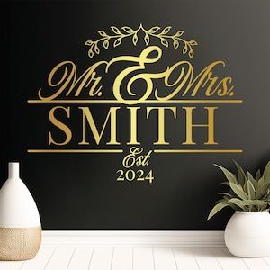 A gold vinyl decal featuring the text 'Mr. & Mrs. SMITH' with decorative elements above and 'Est. 2024' below. The design is set against a dark wall, with a white vase and plant in the foreground, creating an elegant wedding decor look.
