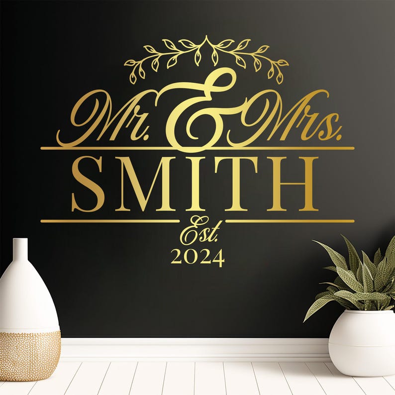 A gold vinyl decal featuring the text 'Mr. & Mrs. SMITH' with decorative elements above and 'Est. 2024' below. The design is set against a dark wall, with a white vase and plant in the foreground, creating an elegant wedding decor look.