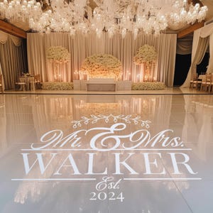 A stylish wedding dance floor featuring a large white vinyl decal that reads 'Mr. & Mrs. WALKER Est. 2024.' The backdrop includes elegant floral arrangements and soft drapery, illuminated by sparkling chandeliers.