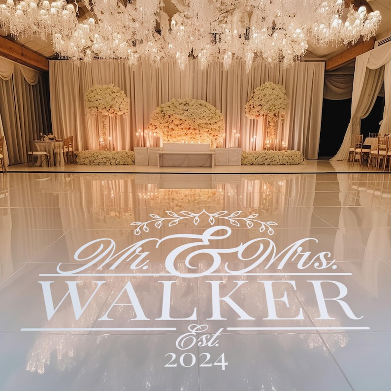 A stylish wedding dance floor featuring a large white vinyl decal that reads 'Mr. & Mrs. WALKER Est. 2024.' The backdrop includes elegant floral arrangements and soft drapery, illuminated by sparkling chandeliers.