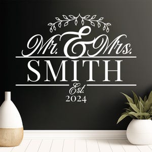 A white vinyl decal featuring the text 'Mr. & Mrs. SMITH' with decorative elements above and the year 'Est. 2024' below. The design is displayed on a dark wall, accompanied by a modern vase and a potted plant.