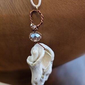 May include: A handmade seashell pendant necklace. The pendant is a natural, off-white seashell, accented with a copper-colored wire wrap and a clear, faceted bead. The necklace cord is a thin, white, woven material.
