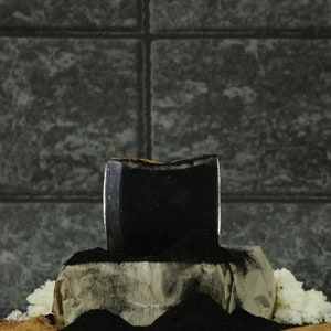 Puede incluir: Un bloque rectangular negro de carbón vegetal se asienta sobre un montón de ceniza gris y negra. El carbón vegetal está rodeado por una superficie blanca texturizada.