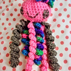 May include: A pink crocheted octopus with a purple and blue bow on its head. The octopus has grey, pink, blue, purple, and green tentacles.