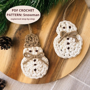 May include: Two handmade crochet snowman ornaments. The snowmen are white with brown accents, including a hat, bow, and button details. The ornaments are hanging on a wooden surface with pine needles and pine cones.