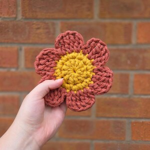 May include: A handmade crochet flower with a yellow center and rust-colored petals. The flower is held in a hand against a brick wall background. The crochet stitches are visible, giving the flower a textured appearance.