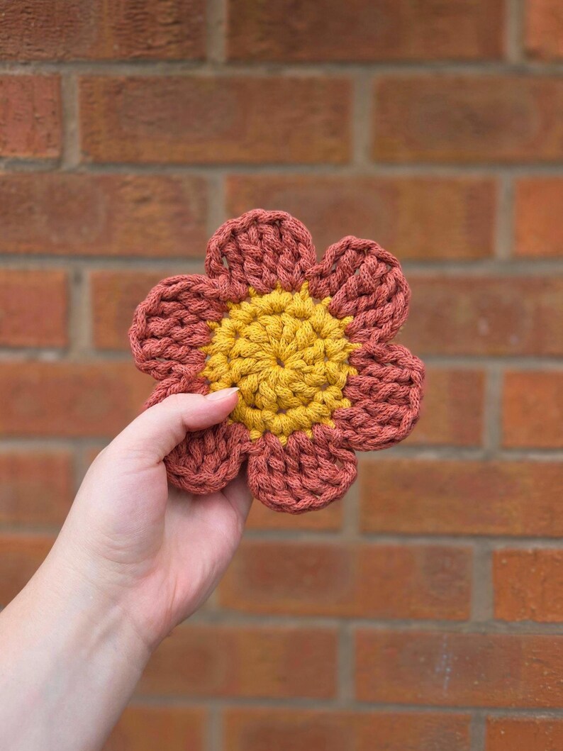 May include: A handmade crochet flower with a yellow center and rust-colored petals. The flower is held in a hand against a brick wall background. The crochet stitches are visible, giving the flower a textured appearance.