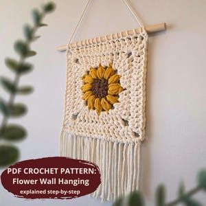 May include: Cream-colored crochet wall hanging with a sunflower design. The sunflower has a brown center and yellow petals. The wall hanging is square with a fringe at the bottom and is suspended from a wooden dowel.