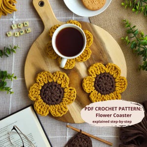 Può includere: Un tagliere in legno mostra una tazza bianca piena di liquido scuro, appoggiata su un sottobicchiere a forma di fiore all'uncinetto giallo e marrone. Sono visibili altri due sottobicchieri. Il testo "TEA TIME" è scritto con tessere. L'immagine include anche il testo "PDF CROCHET PATTERN: Flower Coaster explained step-by-step."