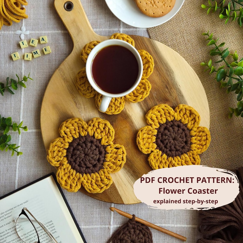May include: A wooden cutting board holds a white teacup filled with dark liquid, resting on a yellow and brown crocheted flower coaster. Two additional coasters are nearby. The text "TEA TIME" is spelled out with letter tiles. The image also includes the text "PDF CROCHET PATTERN: Flower Coaster explained step-by-step."