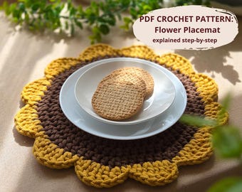May include: A handmade, flower-shaped crochet placemat in brown and yellow. Two white plates hold two round cookies. The placemat has a scalloped edge and is on a light brown surface. Text on the image reads "PDF Crochet Pattern: Flower Placemat explained step-by-step."