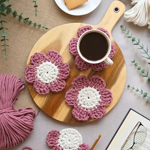 May include: A wooden serving board with a cup of coffee, crackers, and four handmade crochet flower coasters. The coasters are pink with white centers. A ball of pink yarn, a crochet hook, and a book with glasses are also present.