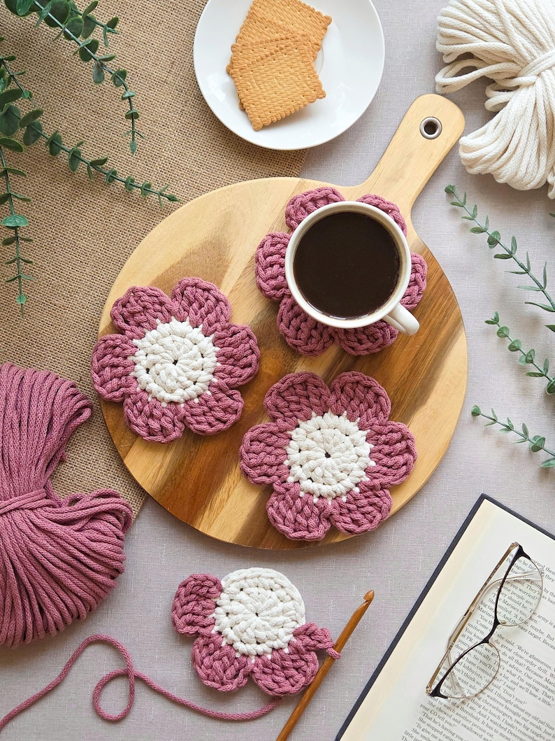 May include: A wooden serving board with a cup of coffee, crackers, and four handmade crochet flower coasters. The coasters are pink with white centers. A ball of pink yarn, a crochet hook, and a book with glasses are also present.
