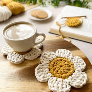 May include: Two handmade, daisy-shaped crochet coasters. The coasters have a white petal design with a yellow center. A white mug of coffee sits on one coaster. The coasters are on a wooden surface.