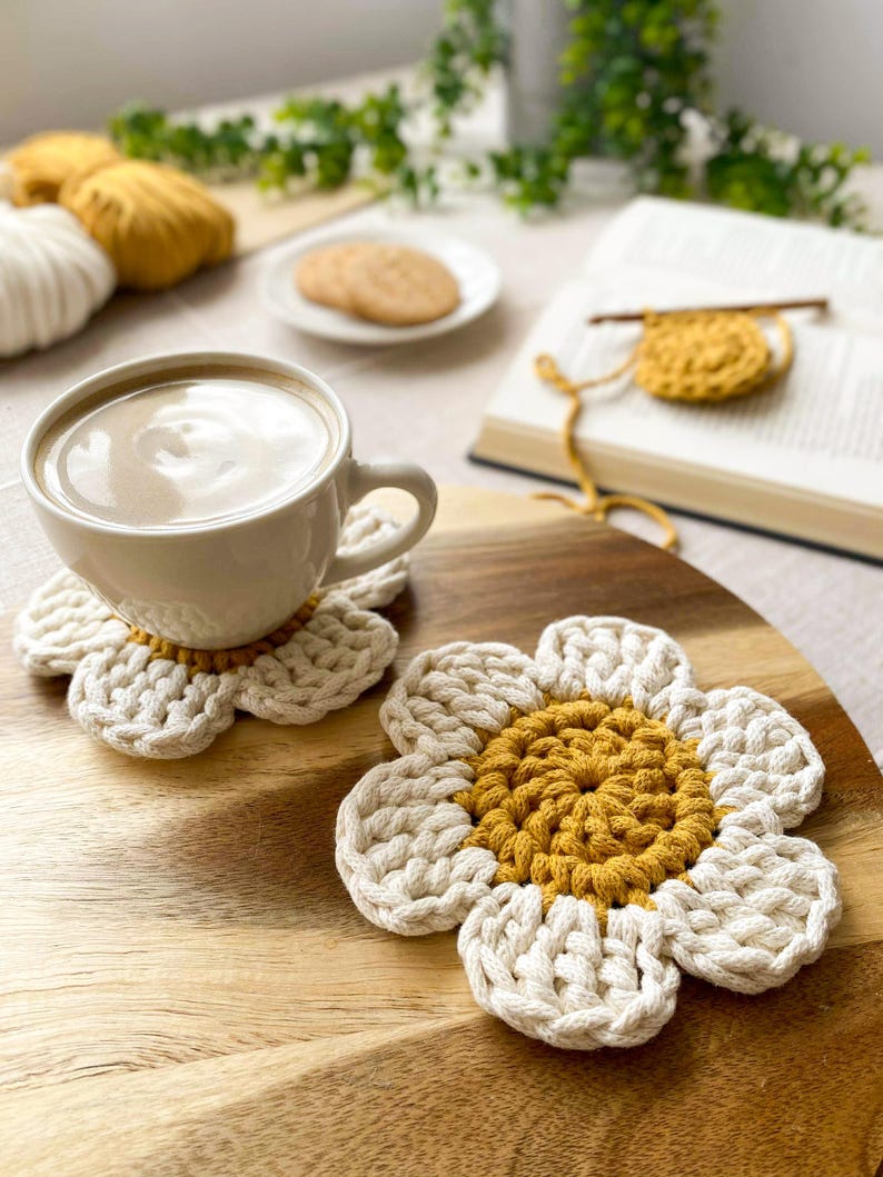 May include: Two handmade, daisy-shaped crochet coasters. The coasters have a white petal design with a yellow center. A white mug of coffee sits on one coaster. The coasters are on a wooden surface.