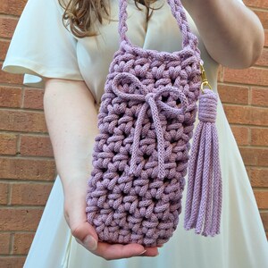 May include: A lilac-coloured, hand-crocheted bag with a plaited strap and a matching tassel. The bag has a decorative bow and a gold-coloured clasp. The bag is designed to hold a water bottle or other small items.