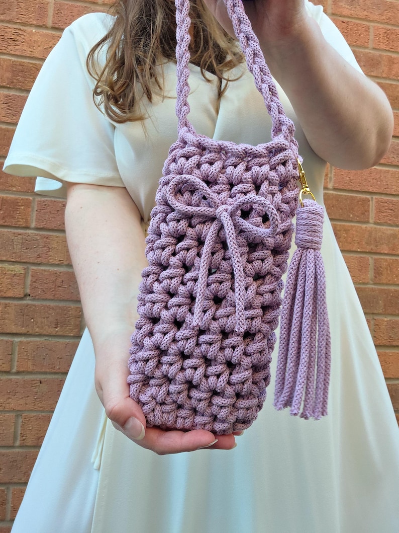May include: A lilac-coloured, hand-crocheted bag with a plaited strap and a matching tassel. The bag has a decorative bow and a gold-coloured clasp. The bag is designed to hold a water bottle or other small items.