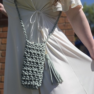 May include: A sage green, hand-crocheted phone crossbody bag with a matching strap and tassel. The bag is made of thick yarn, featuring a textured stitch pattern. The strap is braided, and the tassel hangs from the bottom.