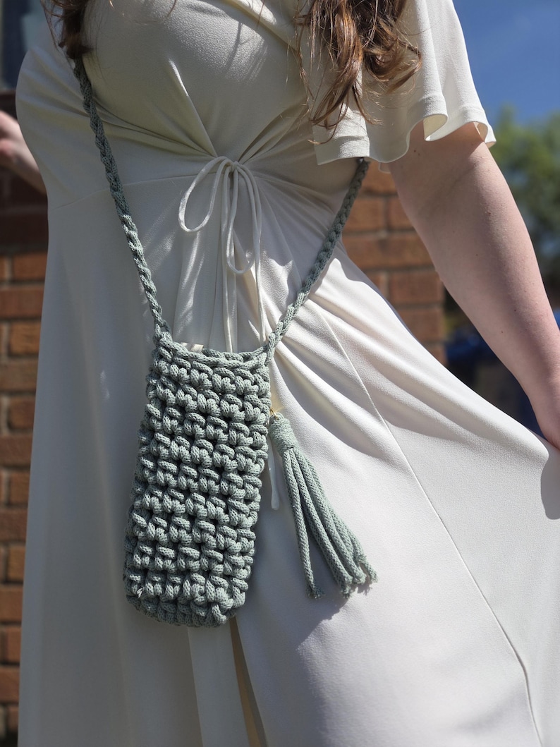 May include: A sage green, hand-crocheted phone crossbody bag with a matching strap and tassel. The bag is made of thick yarn, featuring a textured stitch pattern. The strap is braided, and the tassel hangs from the bottom.