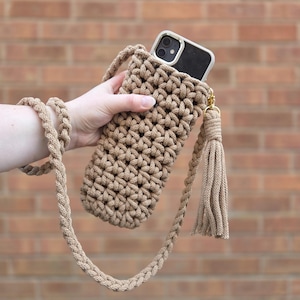 May include: A tan, hand-crocheted phone crossbody bag with a matching braided strap and tassel. The bag holds a black smartphone with a white case. The background is a brick wall.
