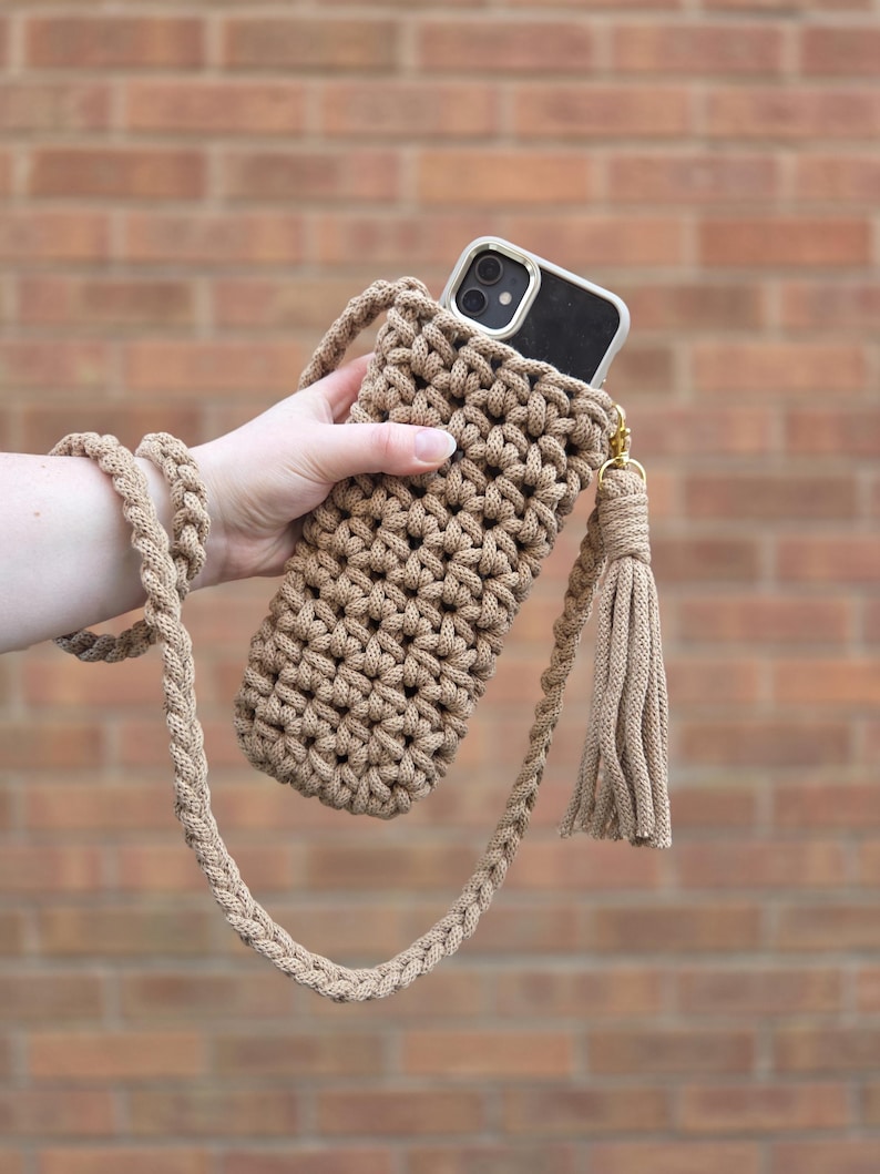 May include: A tan, hand-crocheted phone crossbody bag with a matching braided strap and tassel. The bag holds a black smartphone with a white case. The background is a brick wall.