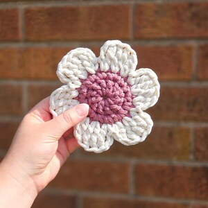May include: A handmade crochet flower coaster with a white petal design and a mauve center. The coaster is held in a hand against a brick background. The flower design is a charming addition to any home decor.