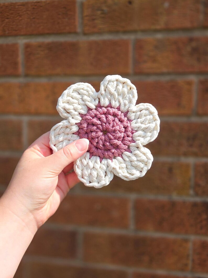 May include: A handmade crochet flower coaster with a white petal design and a mauve center. The coaster is held in a hand against a brick background. The flower design is a charming addition to any home decor.