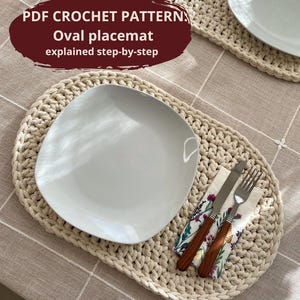 May include: A beige, oval crochet placemat with a white plate, a napkin, and cutlery. The napkin features a floral design. Text on the image reads "PDF CROCHET PATTERN Oval placemat explained step-by-step."