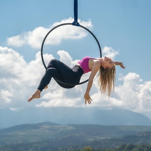May include: A woman in a pink top and black leggings performs aerial acrobatics on a black hoop suspended from a blue strap. The background features a blue sky with white clouds and a mountain range.