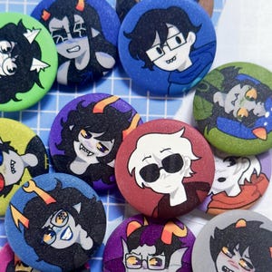 Puede incluir: Una colección de botones redondos y coloridos con ilustraciones de personajes de dibujos animados. Los botones tienen varios colores de fondo, incluyendo azul, verde, morado y rojo. Los personajes tienen diferentes peinados y expresiones.