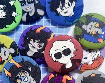 May include: A collection of colorful, round buttons featuring cartoon character illustrations. The buttons have various background colors, including blue, green, purple, and red. The characters have different hair colors and expressions, with some wearing glasses.