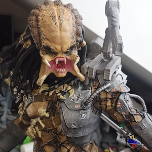 Pode incluir: Um close-up de uma figura de ação do Predator. A figura está vestindo um traje de camuflagem preto e marrom com uma placa de peito prateada e preta. O Predator tem uma cabeça grande e detalhada com uma expressão ameaçadora e dentes afiados. A figura está segurando uma arma em sua mão direita.
