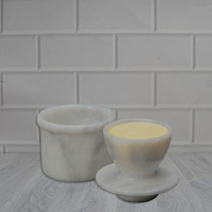 May include: White marble butter dish with a separate lid and a small, round, white marble butter dish with a yellow butter pat inside.